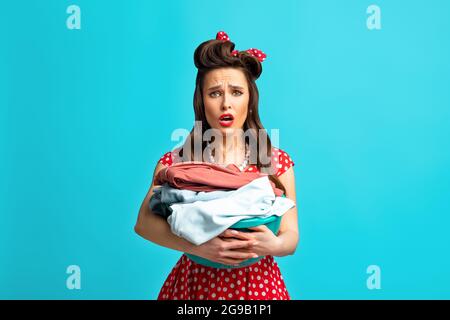 Infelice giovane pinup stanco in abito retrò tenendo bacino con bucato per il lavaggio o la stiratura su sfondo blu Foto Stock