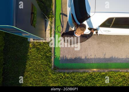 Vista dall'alto di un giovane caucasico che carica un'auto elettrica bianca presso una stazione di ricarica pubblica Foto Stock