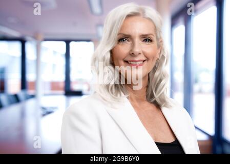 Ritratto di una donna d'affari con capelli grigi che guarda la macchina fotografica e sorride mentre si trova in piedi nella sala riunioni. Spazio di copia. Foto Stock