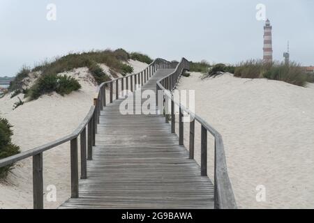 Passerelle in legno lungo le dune della spiaggia di barra con il faro di Aveiro sullo sfondo in una giornata nuvolosa. Portogallo Foto Stock