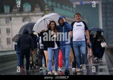 Un gruppo di persone cammina sul Westminster Bridge mentre la pioggia batte il centro di Londra. Tempeste di tuoni che portano fulmini e pioggia torrenziale a sud sono fissati per continuare fino a Lunedi, hanno detto i previsori. Data immagine: Domenica 25 luglio 2021. Foto Stock