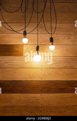 Una lampada composta da tre moderne lampade a LED su lunghi fili neri contro una parete di tavole di legno. Illuminazione moderna ed elegante con lampade retrò su legno d'epoca Foto Stock