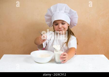 Una bambina vestita come cuoco impasta l'impasto. Cucina bambino stile di vita concetto. Il capretto ama, ha divertimento, studia e gioca nella cucina Foto Stock