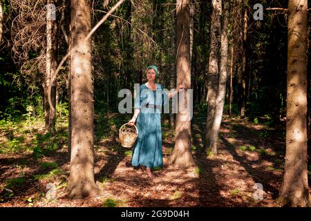 giovane donna in abiti popolari contadini con un cesto di vimini per raccogliere alcune piante selvatiche, bacche o funghi nella foresta di conifere Foto Stock