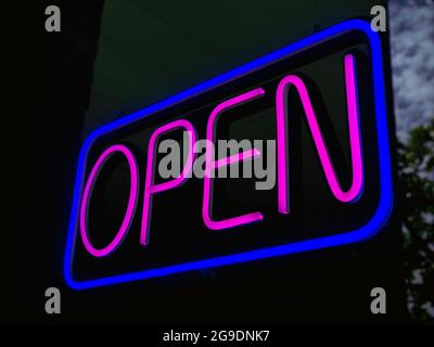 APRI il cartello al neon sul vetro all'entrata del bar o del bar, sfondo scuro, testo rosa illuminato in un cartello quadrato blu che dice 'APERTO', per un cartello d'affari. Foto Stock