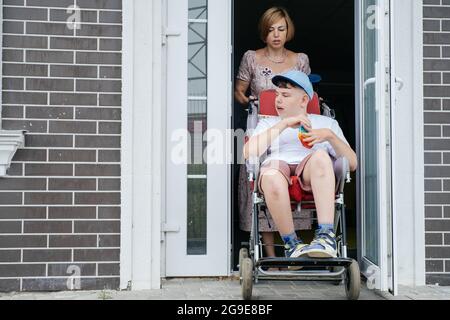 Madre con bambino adolescente disabile in passeggino grande. Accessibilità delle persone in carrozzina. Donna che passa attraverso la porta, persone con bisogni speciali Foto Stock