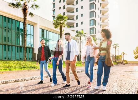 Gruppo multiculturale variegato di giovani amici che camminano in fila lungo una strada urbana chiacchierando e ridendo retroilluminati dal sole in una vista ad angolo basso Foto Stock