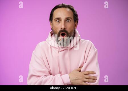 Scioccato impressionato gossiping caucasico bearded adulto 40s uomo capelli grigi con una felpa con cappuccio rosa che spazza le labbra ripiegate affascinate, si allargano gli occhi udito stupefatto Foto Stock