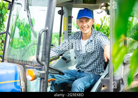 Uomo allegro agricoltore che guida piccolo trattore in frutteto Foto Stock