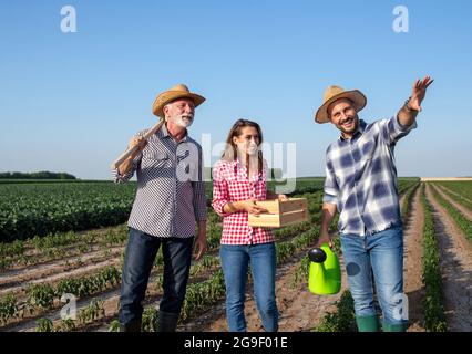 Giovane agricoltore maschio che tiene annaffiatura può indicare mostrando con la mano sollevata. Agricoltore anziano che tiene la zappa da giardinaggio sulla spalla e giovane donna che tiene il woo Foto Stock