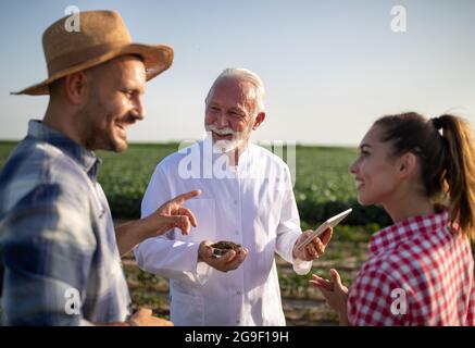 Coppia giovani agricoltori che discutono la qualità del suolo in piedi in campo sorridente. Anziano scienziato che indossa camice da laboratorio utilizzando compressa e piastra Petri con soi Foto Stock