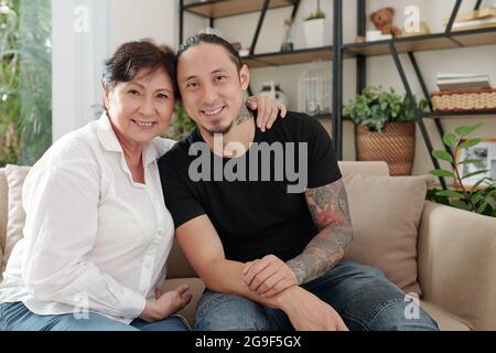 Ritratto di felice madre asiatica abbracciando il suo figlio maggiore mentre si siedono sul divano nella stanza Foto Stock
