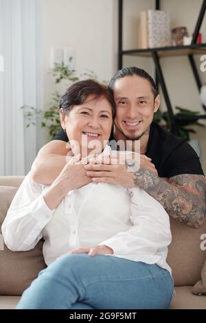 Ritratto della felice famiglia asiatica di due sorridenti con la macchina fotografica mentre si riposa sul divano nella stanza a casa Foto Stock