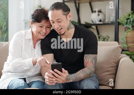 Figlio maggiore che mostra le foto sul suo cellulare alla sua madre felice mentre si riposano sul divano a casa Foto Stock