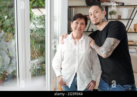 Ritratto di una donna asiatica matura che sorride alla macchina fotografica mentre il suo figlio maggiore la abbracciava nella stanza Foto Stock