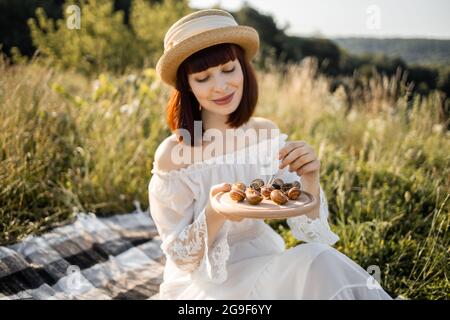 Primo piano ritratto di simpatica giovane donna romantica in abito bianco e cappello di paglia, seduto sul picnic all'aperto e mangiare deliziosi escargot di lumache arrostite. Picnic sulla natura Foto Stock