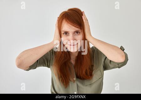 Femmina sorridente con lunghi capelli di zenzero che coprono le orecchie con le mani dal rumore su sfondo bianco e guardando la fotocamera Foto Stock