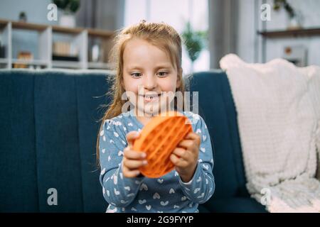 Spingi il gioco di pugnalata pop-it. Pop pit sensoriale. Carino adorabile ragazza ridente eccitata che gioca con il fidget pop arancione, seduto sul divano blu in un accogliente soggiorno. Mettere a fuoco sul viso del bambino Foto Stock