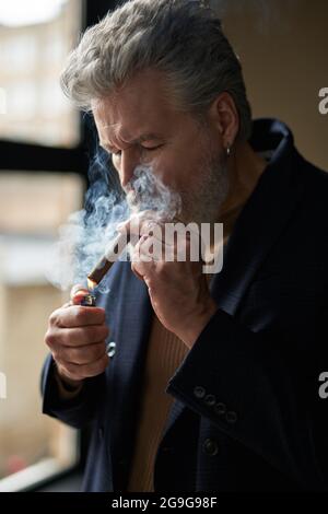 Ritratto di un uomo maturo dai capelli grigi che illumina il sigaro mentre si trova in piedi all'interno Foto Stock