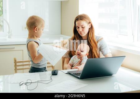 Giovane bella madre con due bambini seduti al tavolo in cucina a casa e lavora su un computer portatile mentre si prende cura dei bambini. Foto Stock