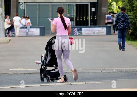 Donna sottile in leggings con un passeggino che attraversa la strada della città con uno smartphone in mano. Concetto di maternità, mamma da passeggio con pRAM Foto Stock