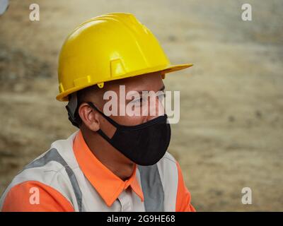 Amaga, Antioquia, Colombia - Luglio 18 2021: Giovane latino indossando una maschera nera, un cappello giallo duro e una divisa da lavoro Foto Stock