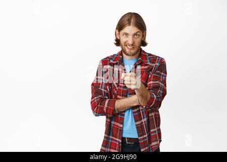 Il ragazzo biondo scettico che punta il dito alla macchina fotografica con incredulità, avendo dubbi, in piedi in vestito casual contro sfondo bianco Foto Stock