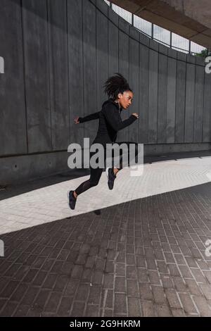 Vista laterale di un corridore afroamericano che salta vicino all'edificio sulla strada urbana Foto Stock