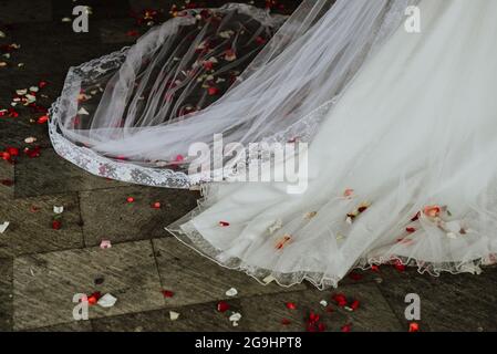 Primo piano dell'abito della sposa e del lungo velo sul terreno coperto di petali di rosa Foto Stock