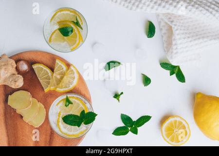 Due bicchieri con limonata e ingredienti per cucinare. Zenzero, limone, menta, ghiaccio su superficie bianca. Vassoio di legno piatto. Vista dall'alto Foto Stock