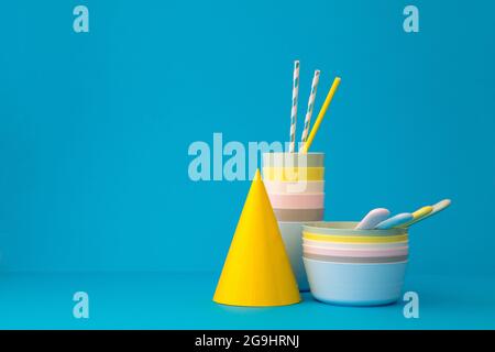 Composizione con stoviglie riutilizzabili in plastica multicolore con cannucce e cappello giallo brillante su sfondo blu Foto Stock