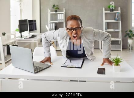 Arrabbiato pazzo stressato donna d'affari nera appoggiata sulla sua scrivania d'ufficio e gridando Foto Stock