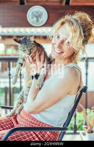 ritratto di una giovane bionda caucasica matura che sorride con il gatto a casa sul portico seduto su una sedia a dondolo. Concetto di stile di vita. Foto Stock