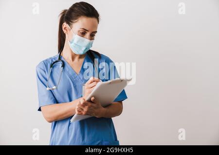 Giovane donna sicura specialista medico indossando uniforme tenendo blocco note isolato su sfondo bianco, prendendo appunti Foto Stock