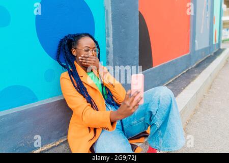 Italia, Milano, Donna con trecce seduta da parete colorata, utilizzando smartphone Foto Stock