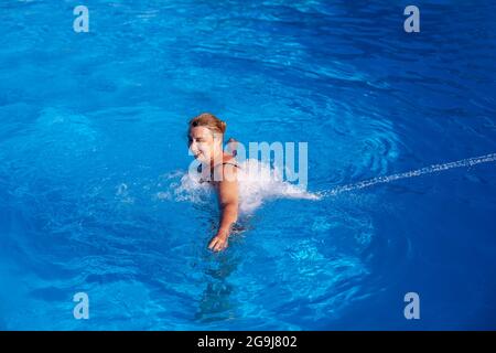 Una bella donna caucasica gode di un idromassaggio alla schiena e al collo con un getto d'acqua in una piscina all'aperto in vacanza Foto Stock