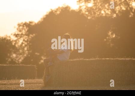 ragazza adolescente con lunghi capelli biondi seduta su una balla di paglia in luce solare calda. Immagine orizzontale di una scena rurale Foto Stock