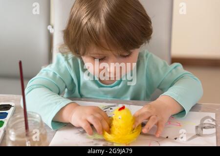 Una ragazza di tre anni in una blusa turchese con entusiasmo e con un sorriso fa mestieri-dipinge un pollo giallo. Vista dall'alto Foto Stock