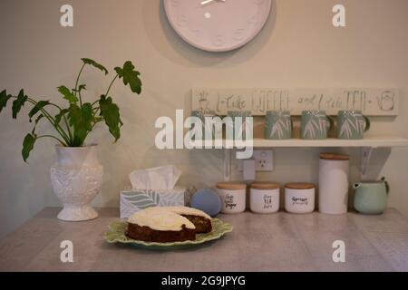 Panca dell'isola della cucina con le tazze del caffè, orologio, altoparlante di Alexa, pianta della casa e torta ghiacciata Foto Stock