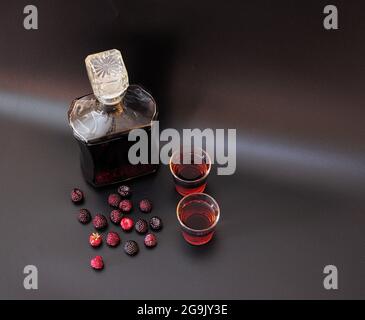 Liquore di mora fatto in casa in una bottiglia e due bicchieri su sfondo nero accanto alle bacche mature. Vista dall'alto. Foto Stock
