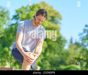 Giovane uomo preoccupato in abiti sportivi che soffrono di dolore al ginocchio mentre corre all'aperto Foto Stock