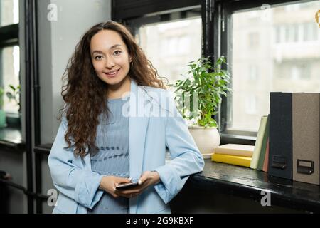 Felice giovane donna d'affari con capelli ondulati scuri lunghi in piedi vicino alla finestra Foto Stock