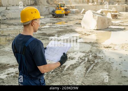 Giovane costruttore maschile in abbigliamento da lavoro che tiene un disegno mentre si trova in cava terra Foto Stock