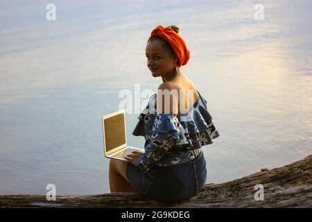 Una giovane e bella donna d'affari malese musulmana con un turbante arancione hijab in testa, che lavora su un computer portatile seduto vicino al mare in una giornata estiva. Lavoro a distanza, l Foto Stock