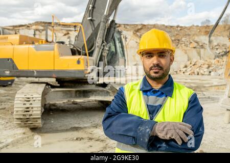Costruttore serio in attrezzatura che attraversa i bracci sul petto contro il bulldozer Foto Stock