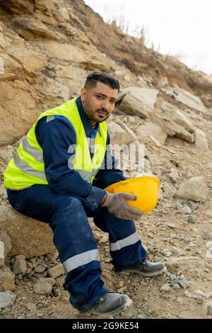 Costruttore di razza mista stanco con cappello in mani seduto da cantiere a pausa Foto Stock