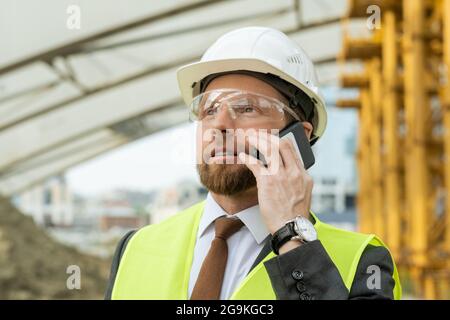 Giovane ingegnere in casco da lavoro che parla sul telefono cellulare mentre si lavora in cantiere Foto Stock