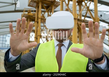 Ritratto di ingegnere in abiti riflettenti in piedi sul cantiere e gesturando lui utilizzando occhiali VR nel suo lavoro Foto Stock
