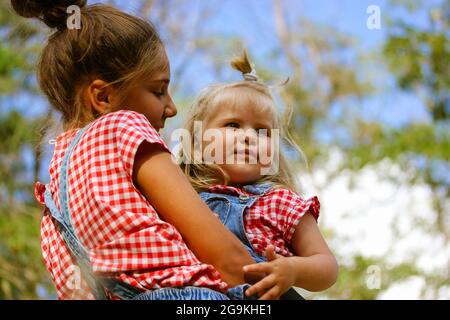 Due sorelle felici nella foresta estiva. Adolescente mestizo ragazza che tiene il bambino sorella bionda ragazza 3 anni. Buona famiglia. Persone correlate. Mostrare amore Foto Stock