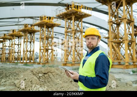 Ritratto di un ingegnere nel casco da lavoro che guarda la fotocamera mentre lavora su un tablet digitale e controlla la costruzione dell'edificio all'aperto Foto Stock